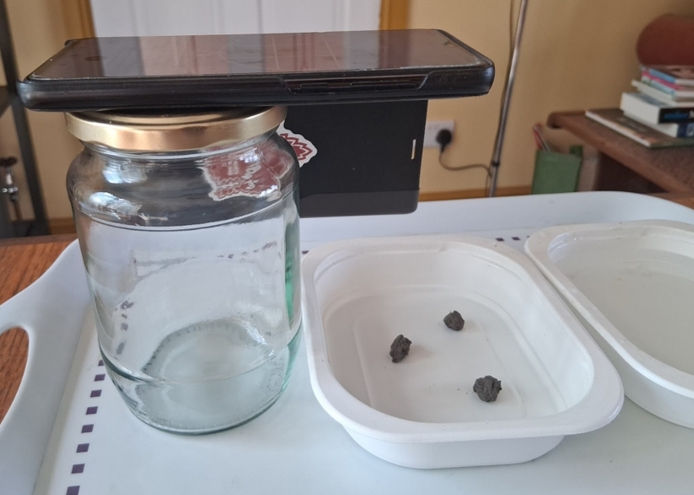 Phone balanced on a jar beside a tray containing soil aggregates, arranged for an at-home test.